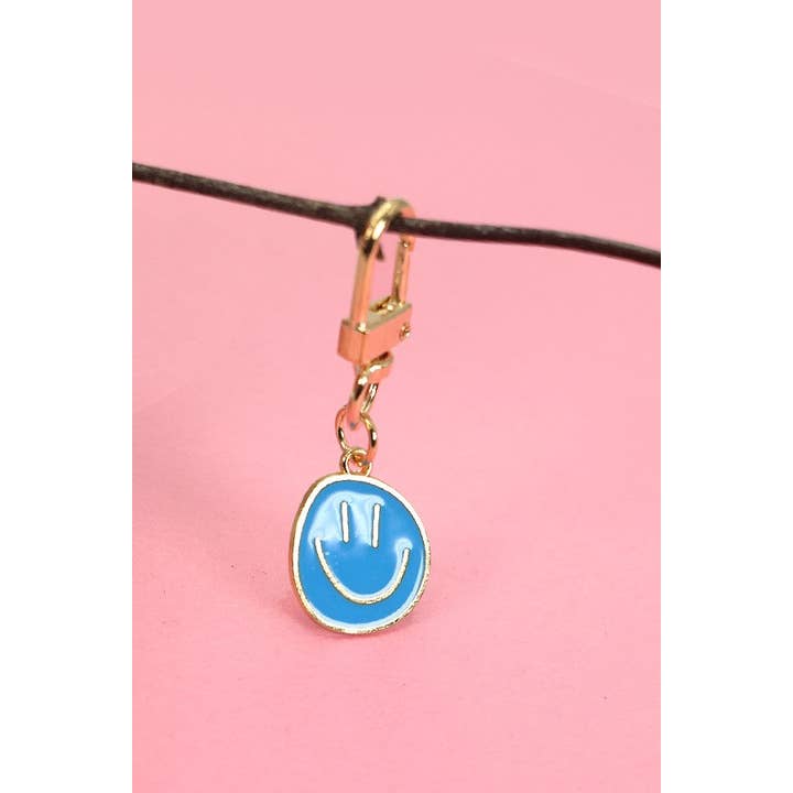 A sky blue keychain with a smiling face design, attached to a gold-colored metal ring, displayed against a pink background.