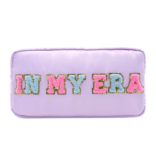A nylon cosmetic bag with the text 'IN MY ERA' embroidered in multicolored letters on a purple background with a gold zipper.