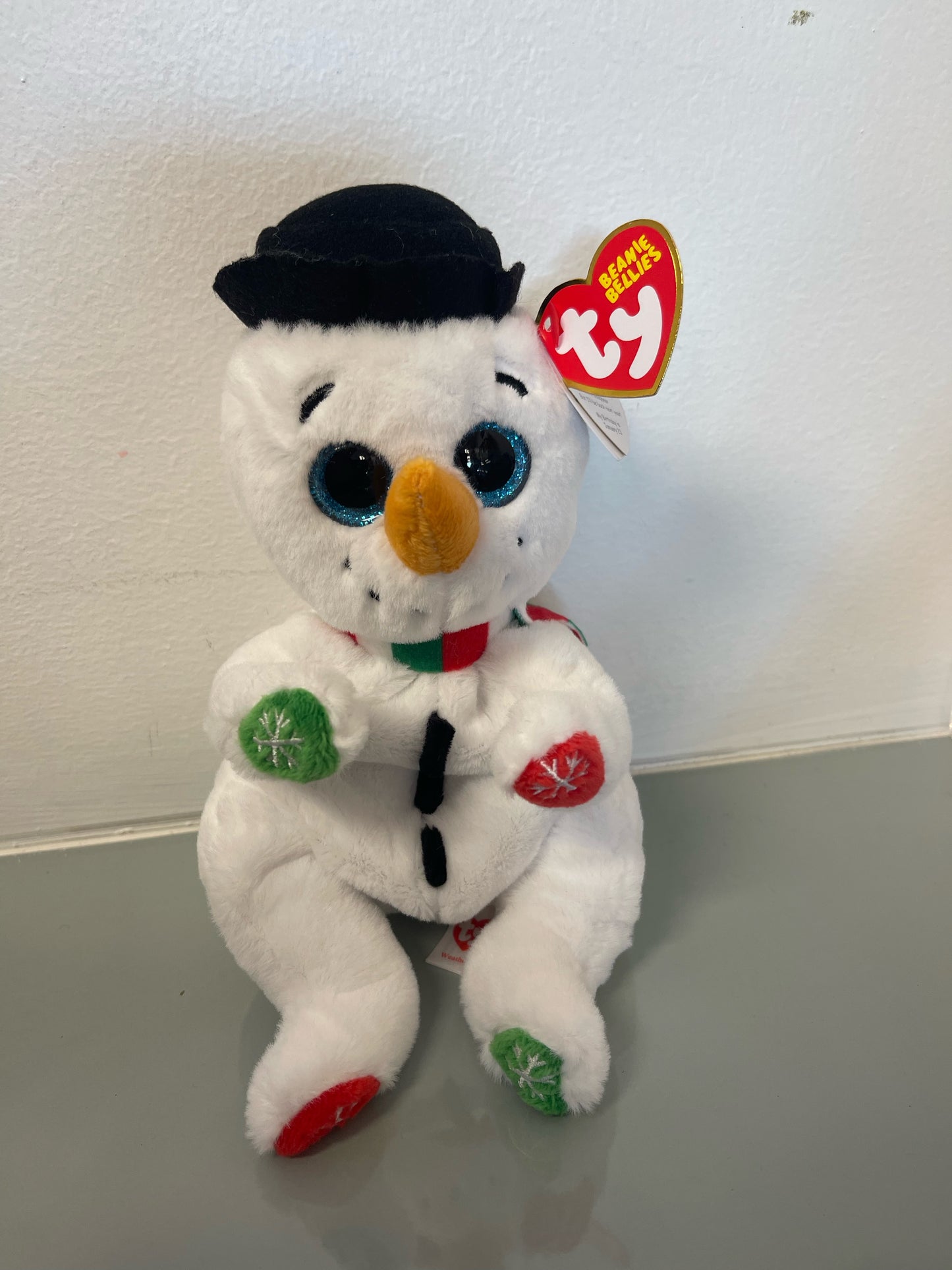 A plush toy resembling a cartoon-style snowman with a green hat and red and green accents on a white body. The toy is marked with a 'Best Toy' sticker from Ty.