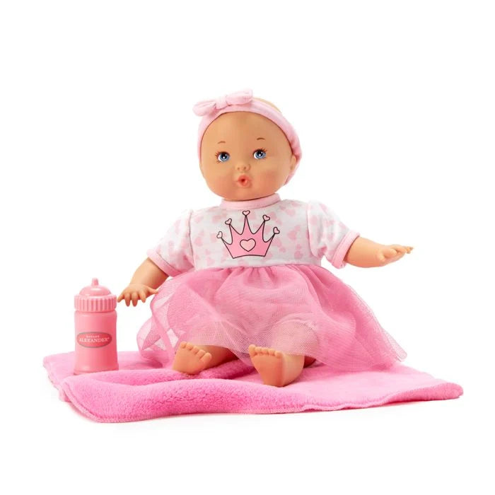 A 12-inch pink baby doll with a soft fabric body, dressed in a pink princess skirt and a top with a printed royal crown, holding a bottle, and placed on a cozy pink blanket.